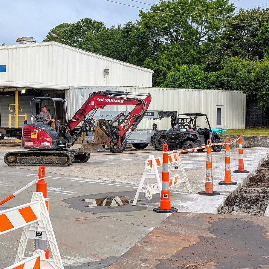 Site work: a mini excavator beside a trench cut through concrete, coned off