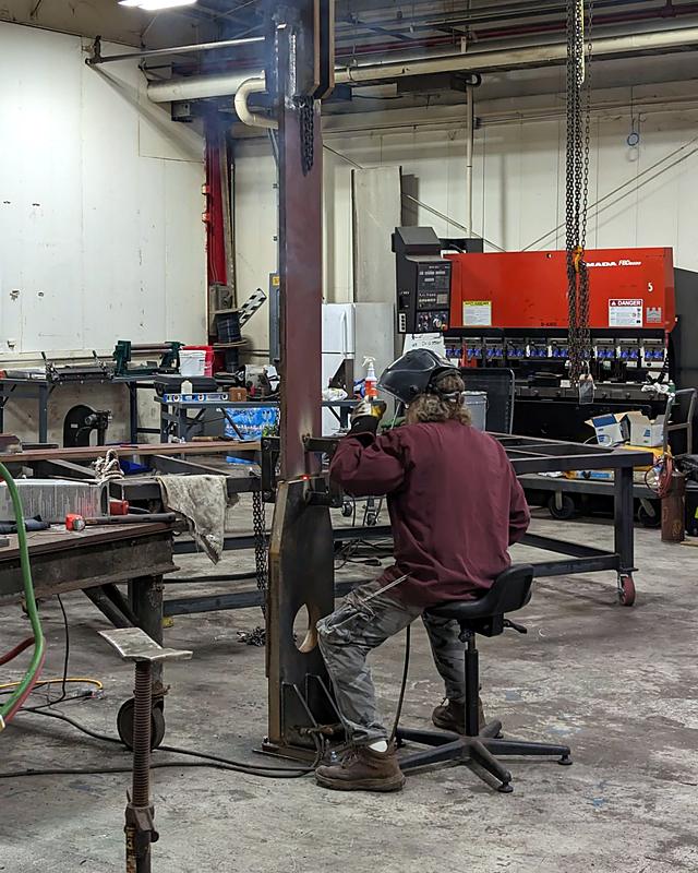 One of the nine lifting adapters being welded while it hangs from a chain hoist, a press brake behind
